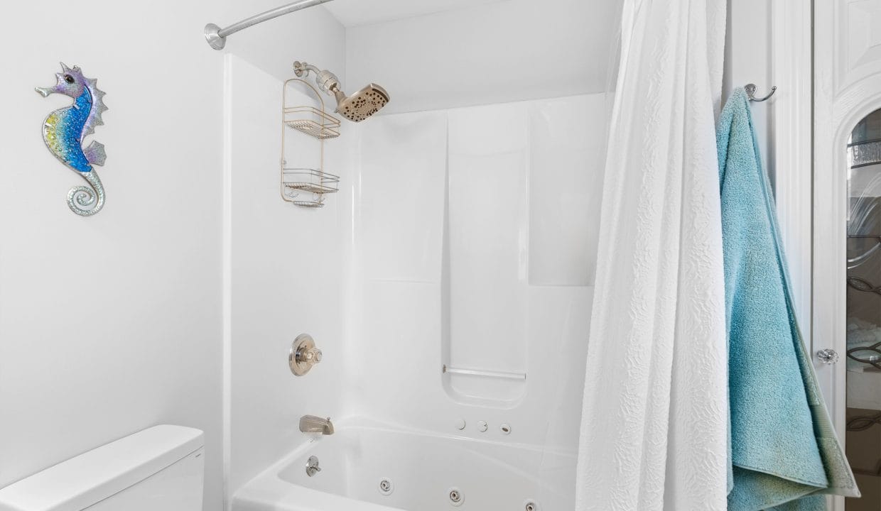 White bathroom with a jetted bathtub and shower, white curtain, blue and green towels, a metal shower caddy, and a decorative seahorse wall ornament above the toilet.