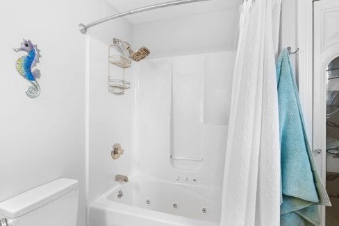 White bathroom with a jetted bathtub and shower, white curtain, blue and green towels, a metal shower caddy, and a decorative seahorse wall ornament above the toilet.