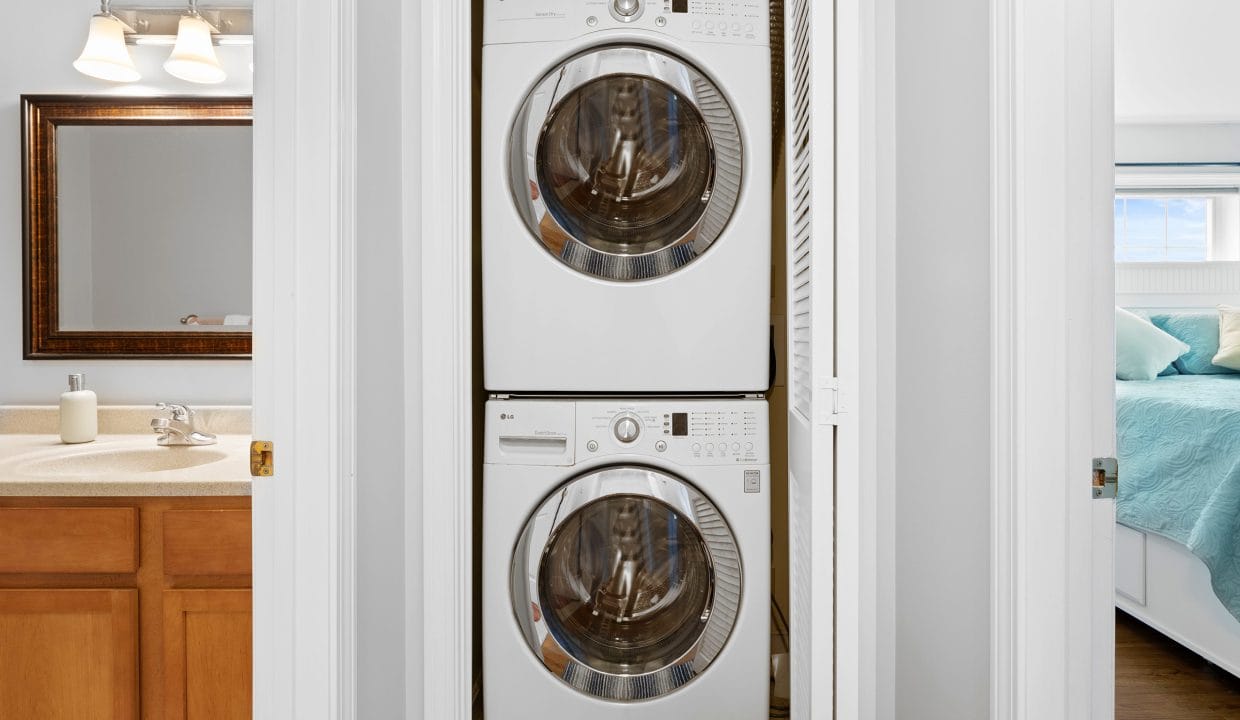 A stacked washer and dryer unit is installed in a closet between a bathroom with a sink and a bedroom with a made bed.