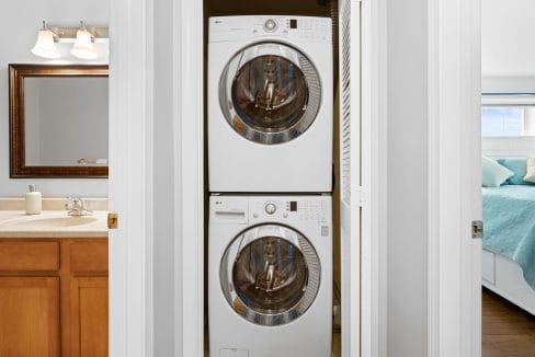 A stacked washer and dryer unit is installed in a closet between a bathroom with a sink and a bedroom with a made bed.