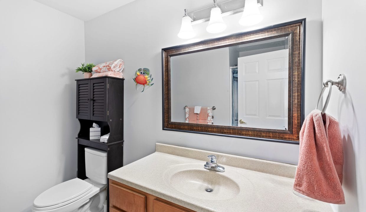 Bathroom with a large mirror above a beige countertop sink, a towel on a hook, a toilet with a dark cabinet, and overhead lighting.