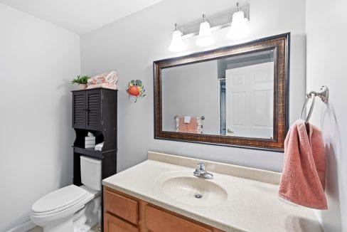 Bathroom with a large mirror above a beige countertop sink, a towel on a hook, a toilet with a dark cabinet, and overhead lighting.