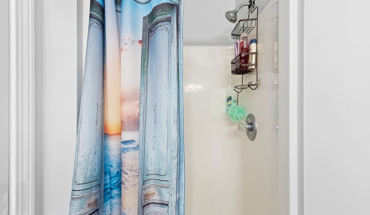 Shower stall with a beach-themed curtain, overhead showerhead, metal shelf holding toiletries, and a green bath sponge hanging from the faucet.