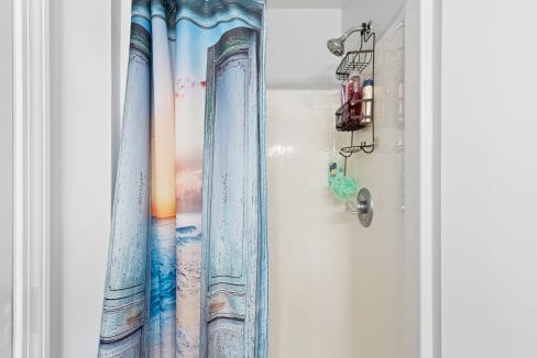 Shower stall with a beach-themed curtain, overhead showerhead, metal shelf holding toiletries, and a green bath sponge hanging from the faucet.
