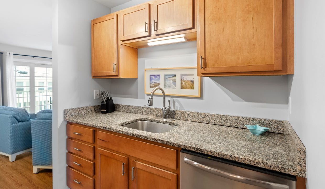 A small kitchen area with wooden cabinets, granite countertops, a stainless steel sink, dishwasher, knife block, and framed pictures on the wall. Blue chair backs are visible in the background.