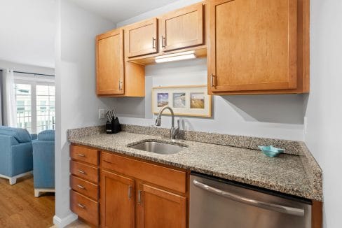 A small kitchen area with wooden cabinets, granite countertops, a stainless steel sink, dishwasher, knife block, and framed pictures on the wall. Blue chair backs are visible in the background.
