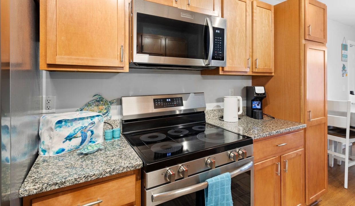 Modern kitchen with stainless steel appliances, light wood cabinets, granite countertops, electric stove, microwave, coffee maker, and a blue towel hanging on the oven handle.