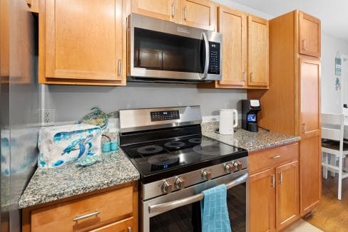 Modern kitchen with stainless steel appliances, light wood cabinets, granite countertops, electric stove, microwave, coffee maker, and a blue towel hanging on the oven handle.