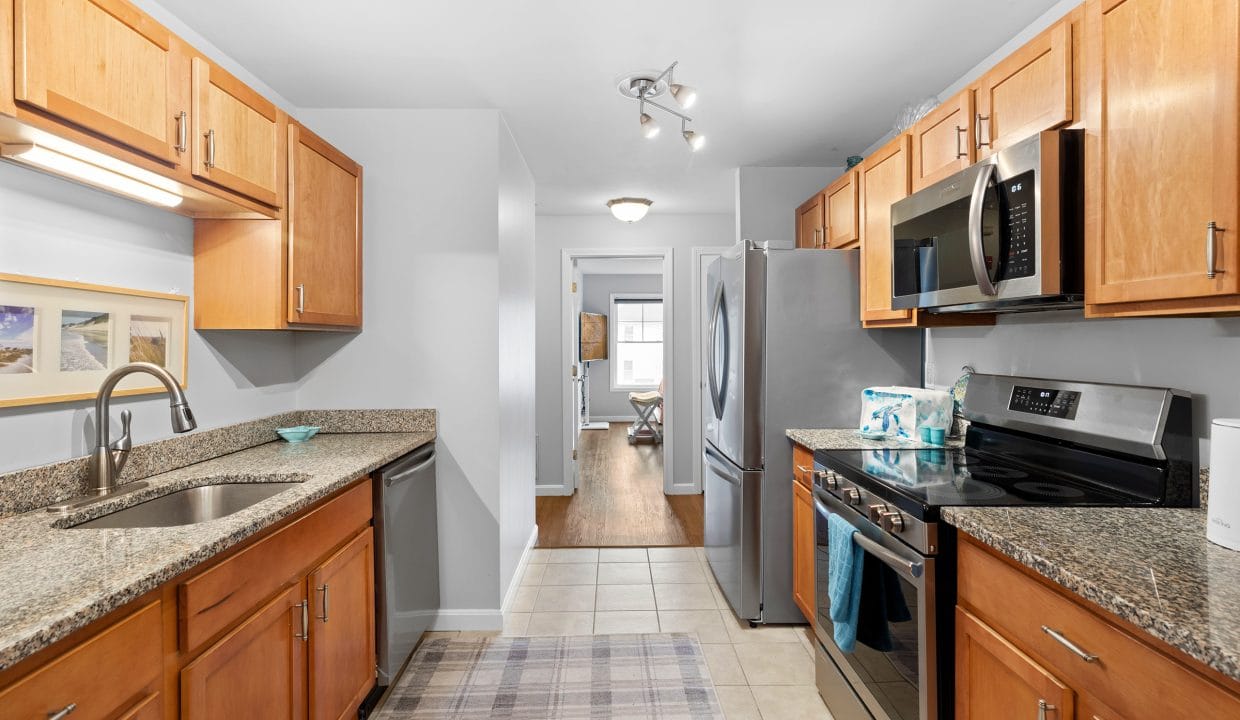 A modern galley kitchen with wood cabinets, granite countertops, stainless steel appliances, and a view into a dining area at the far end.