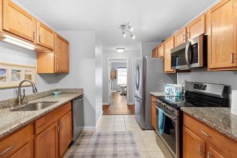 A modern galley kitchen with wood cabinets, granite countertops, stainless steel appliances, and a view into a dining area at the far end.