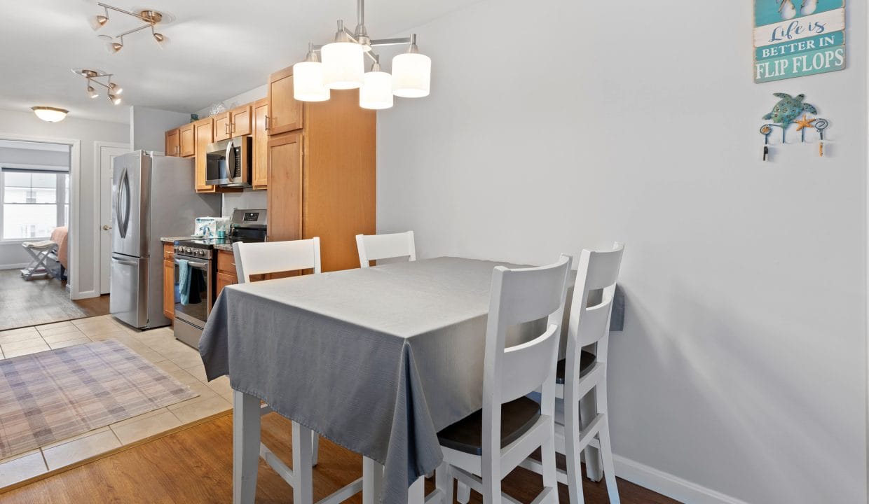 Dining area with a gray tablecloth and four white chairs next to a kitchen with wood cabinets; a decorative sign hangs on the wall.