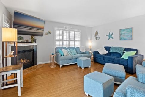 Living room with blue and teal furniture, a fireplace with a TV above it, wooden flooring, and coastal-themed wall decor. Natural light enters through a window with white shutters.