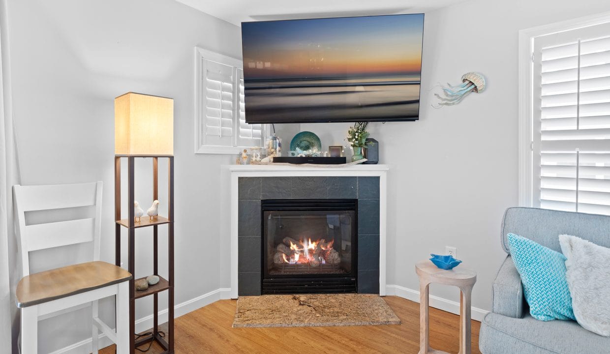 A living room corner with a gas fireplace, a flat-screen TV above, a floor lamp, a chair, a side table, and coastal-themed decor, including a jellyfish wall art piece.