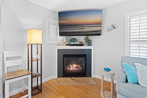 A living room corner with a gas fireplace, a flat-screen TV above, a floor lamp, a chair, a side table, and coastal-themed decor, including a jellyfish wall art piece.