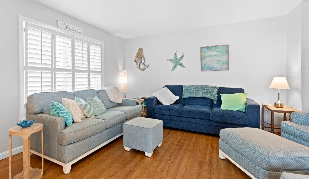 A bright living room with two blue sofas, an ottoman, coastal decor on the walls, a side table with a lamp, and wooden flooring.