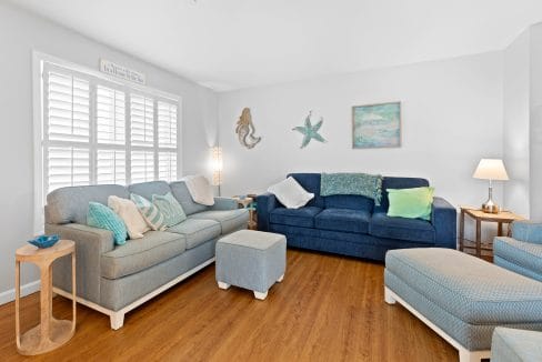 A bright living room with two blue sofas, an ottoman, coastal decor on the walls, a side table with a lamp, and wooden flooring.