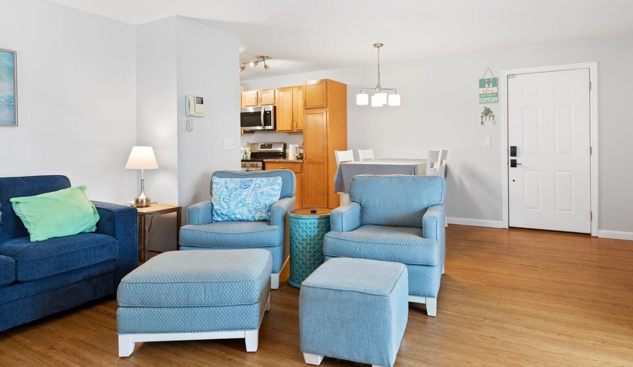 A living room with blue armchairs, ottomans, a side table, and a blue sofa, adjacent to a dining area and kitchen with wooden cabinets and white walls.