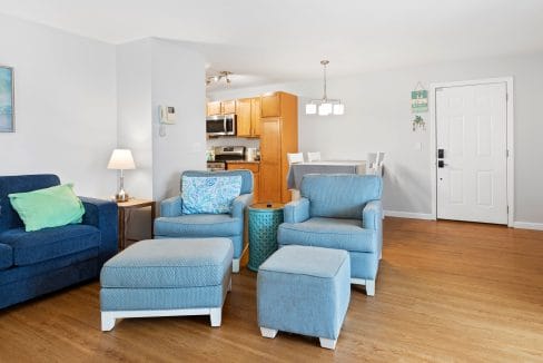 A living room with blue armchairs, ottomans, a side table, and a blue sofa, adjacent to a dining area and kitchen with wooden cabinets and white walls.