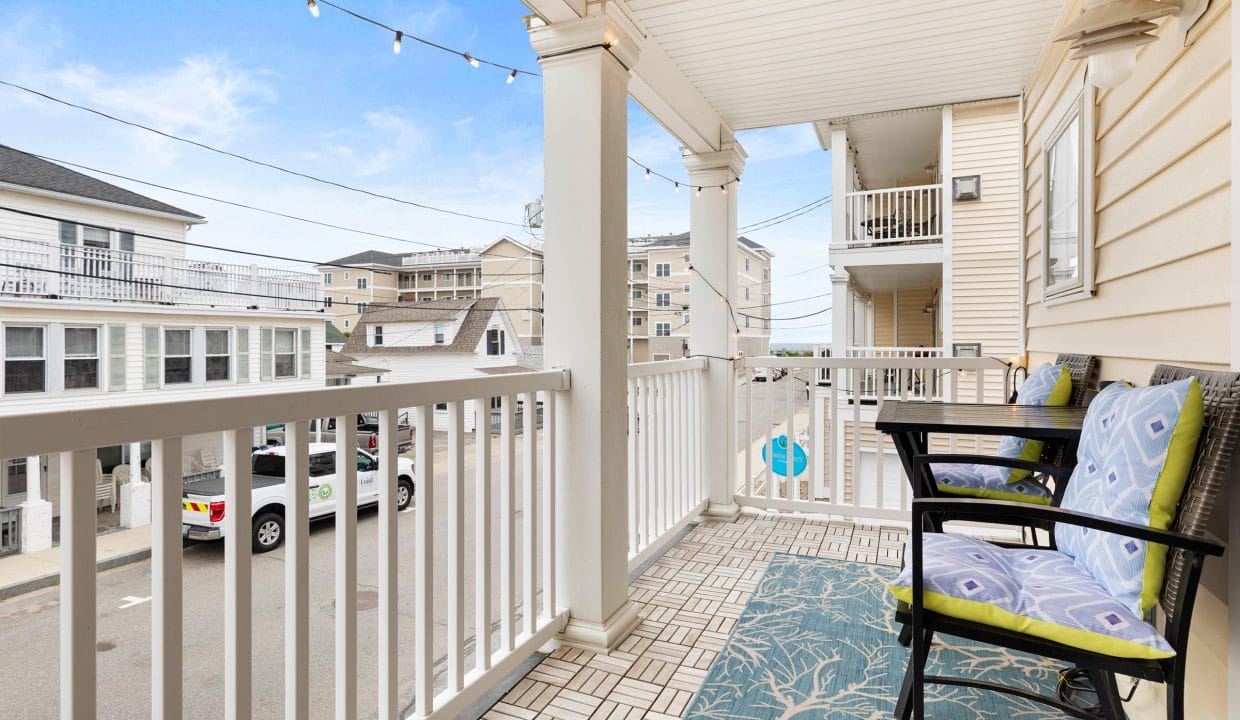 A covered balcony with a patterned rug, black bench with blue cushions, string lights above, and a view of neighboring buildings and parked cars on a clear day.