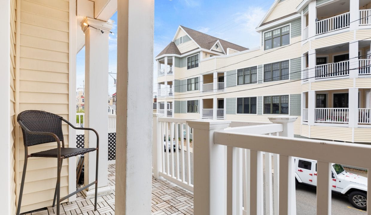 A small balcony with a wicker chair, white railing, and string lights overlooks neighboring multi-story apartment buildings.