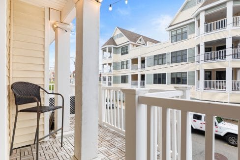 A small balcony with a wicker chair, white railing, and string lights overlooks neighboring multi-story apartment buildings.