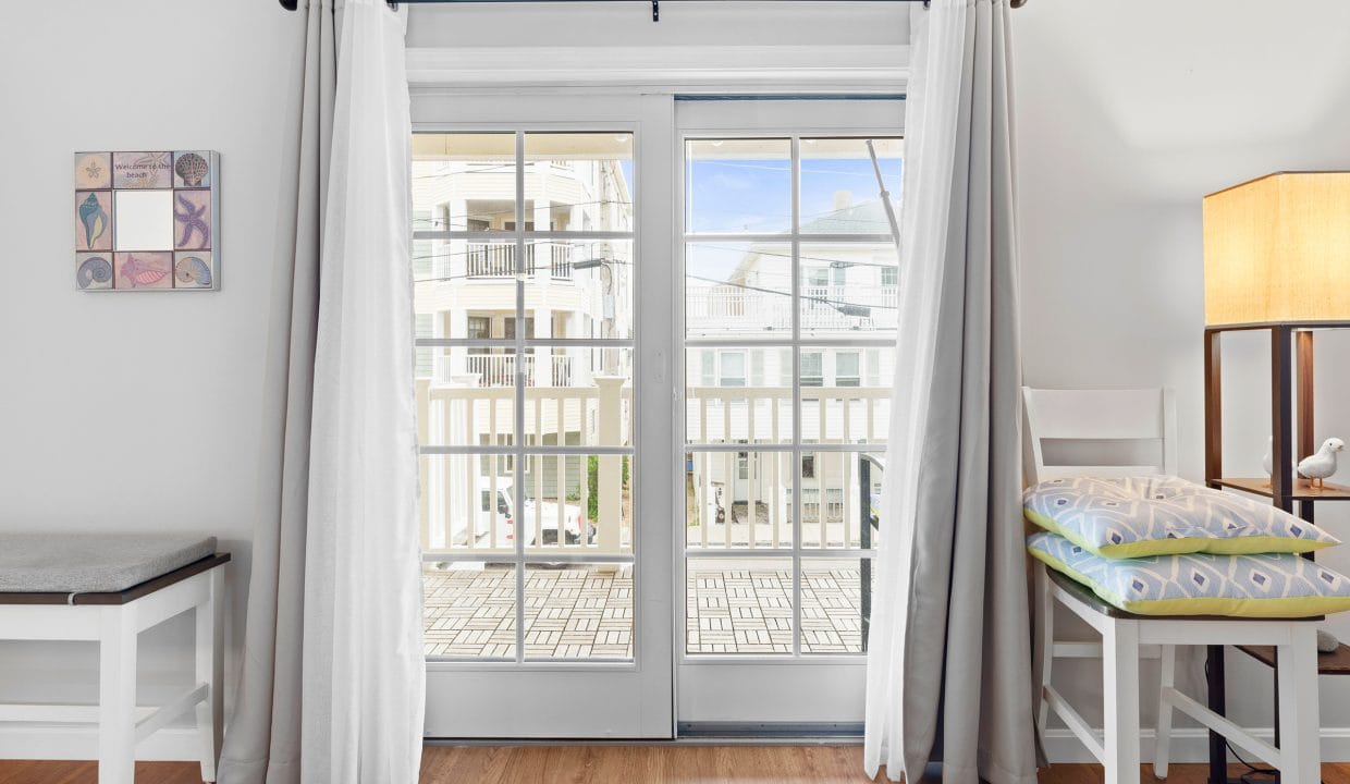 Bright room with wooden floor, a French door with white curtains leading to a balcony, a lamp, a chair with cushions, and wall art on the left.