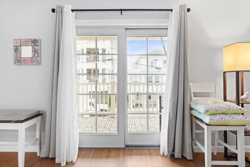 Bright room with wooden floor, a French door with white curtains leading to a balcony, a lamp, a chair with cushions, and wall art on the left.