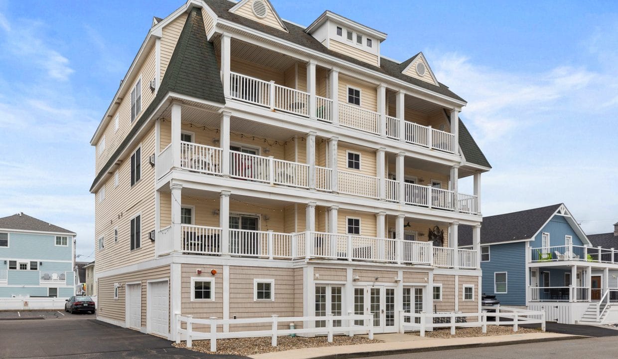 A three-story beige residential building with multiple balconies and white railings, surrounded by a white fence, under a blue sky.