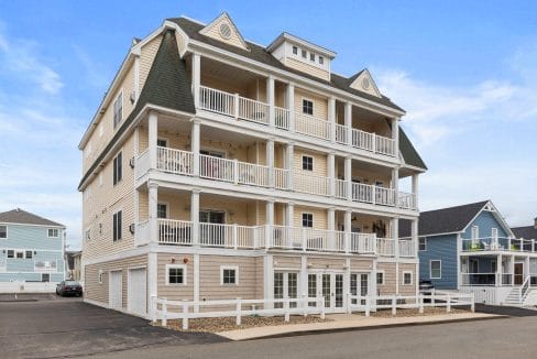 A three-story beige residential building with multiple balconies and white railings, surrounded by a white fence, under a blue sky.