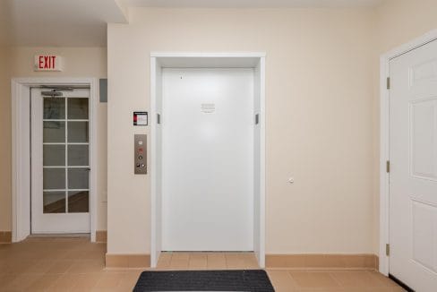 A closed white elevator door is centered in a beige hallway, with an exit sign and a glass door to the left and a solid white door to the right.