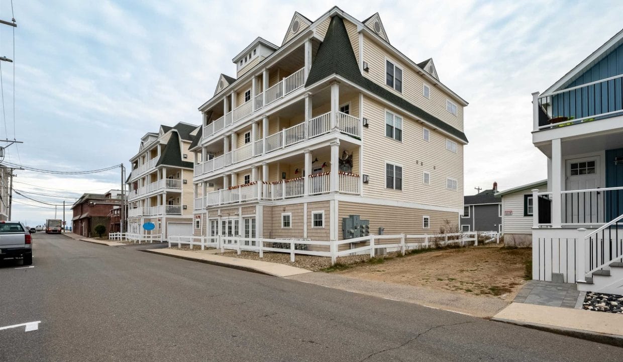 A four-story beige apartment building with multiple balconies is situated on a corner lot along a paved street in a residential neighborhood.