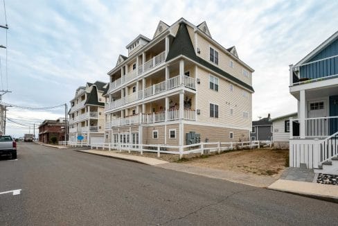 A four-story beige apartment building with multiple balconies is situated on a corner lot along a paved street in a residential neighborhood.