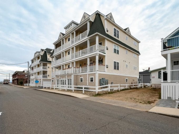 A four-story beige apartment building with multiple balconies is situated on a corner lot along a paved street in a residential neighborhood.