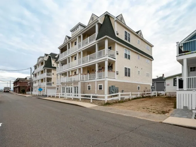 A four-story beige apartment building with multiple balconies is situated on a corner lot along a paved street in a residential neighborhood.