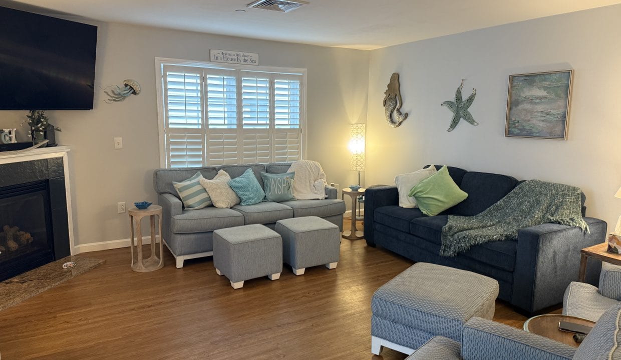 A cozy living room with two gray sofas, two ottomans, a fireplace, wall-mounted TV, and ocean-themed decor on light-colored walls. Wooden floor and a window with white shutters.