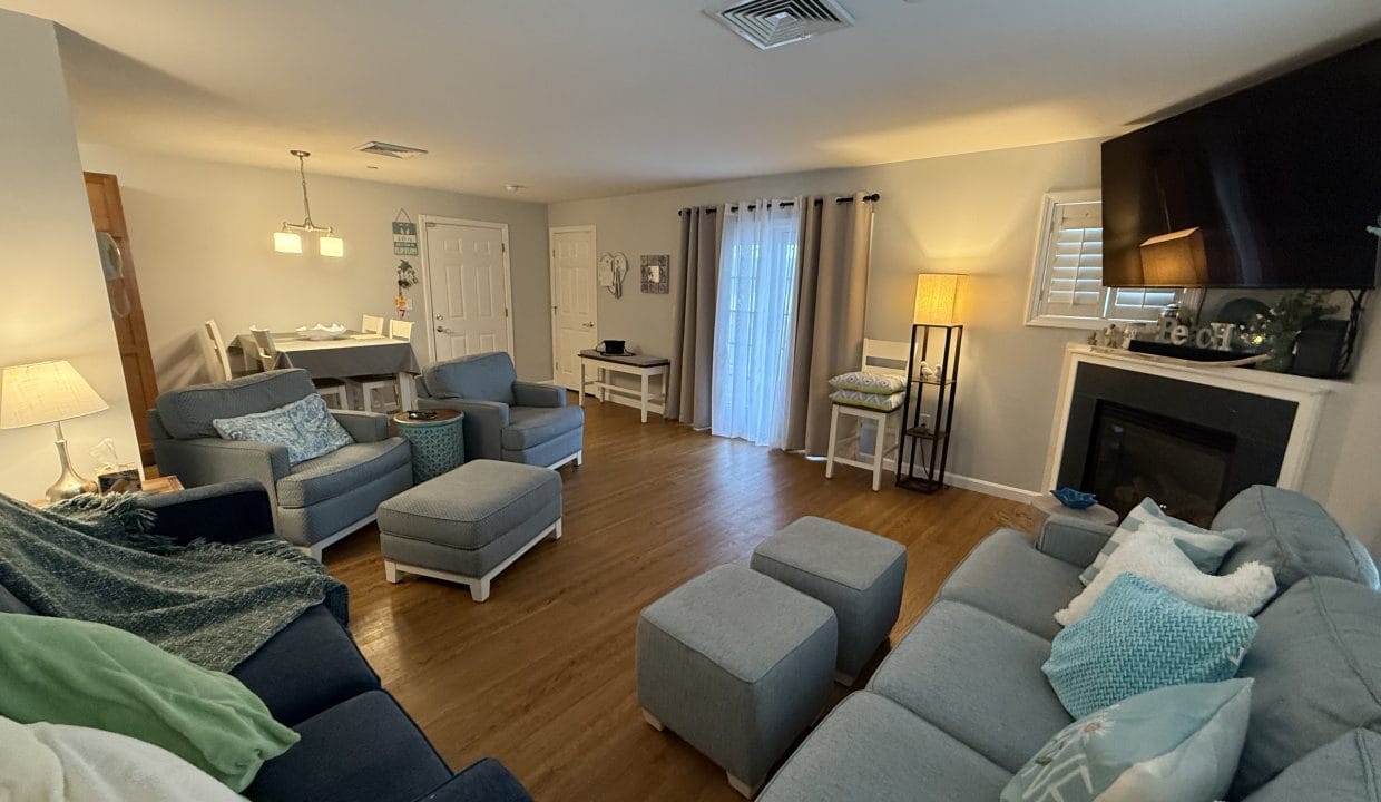 A spacious living room with gray sofas, ottomans, and chairs, a fireplace, a wall-mounted TV, and a dining area with a table and chairs in the background.