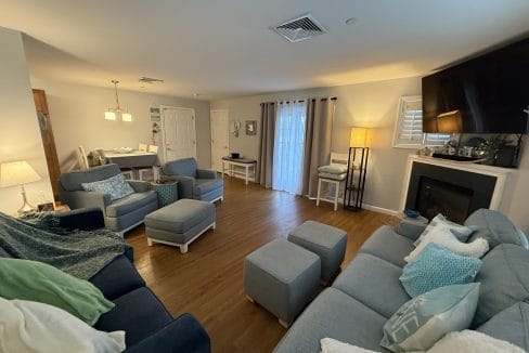 A spacious living room with gray sofas, ottomans, and chairs, a fireplace, a wall-mounted TV, and a dining area with a table and chairs in the background.