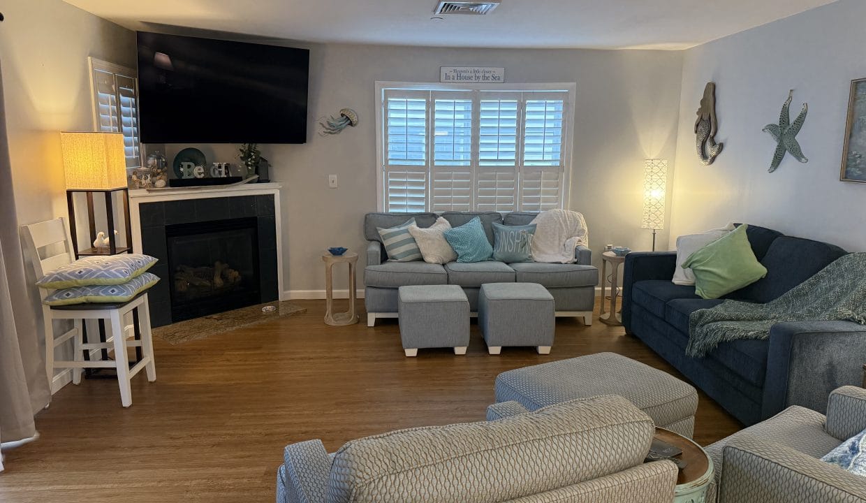 A cozy living room with a sectional sofa, armchairs, ottomans, a wall-mounted TV above a fireplace, and coastal-themed decor on light-colored walls and wood flooring.