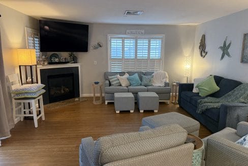 A cozy living room with a sectional sofa, armchairs, ottomans, a wall-mounted TV above a fireplace, and coastal-themed decor on light-colored walls and wood flooring.