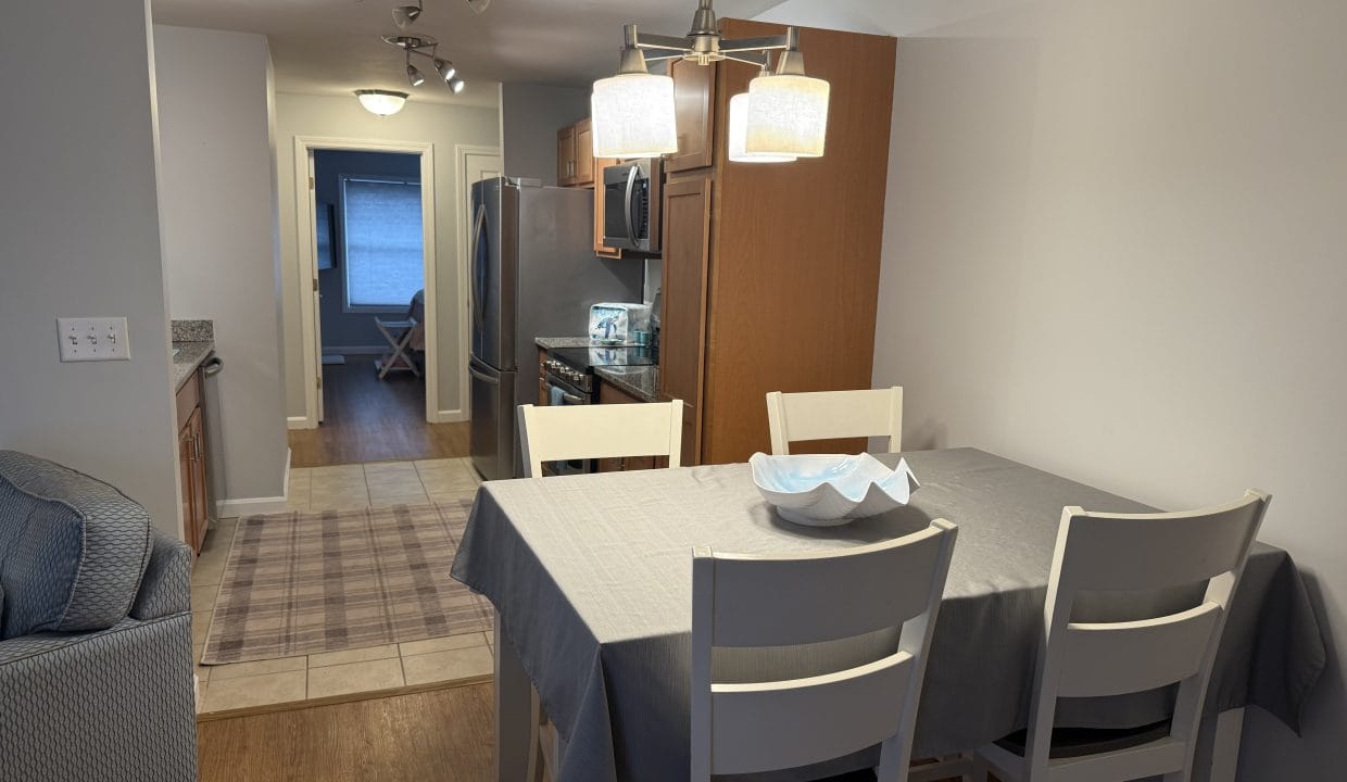 A dining area with a gray tablecloth, four white chairs, and a decorative bowl, adjacent to a kitchen with wooden cabinets and stainless steel appliances.