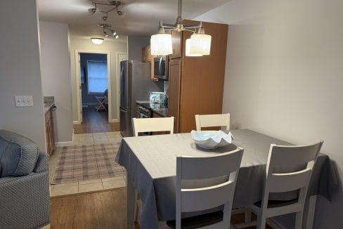 A dining area with a gray tablecloth, four white chairs, and a decorative bowl, adjacent to a kitchen with wooden cabinets and stainless steel appliances.