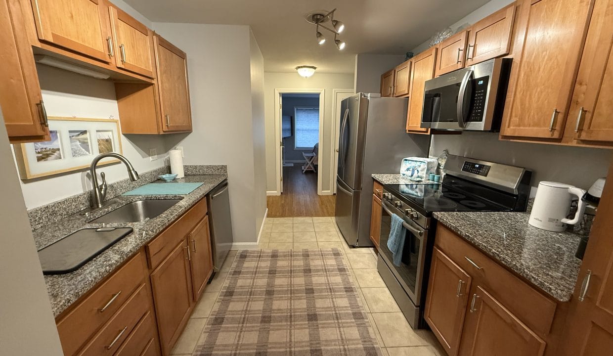 Galley kitchen with wooden cabinets, granite countertops, stainless steel appliances, and a plaid rug on a tiled floor, leading to a dining area with wood flooring.