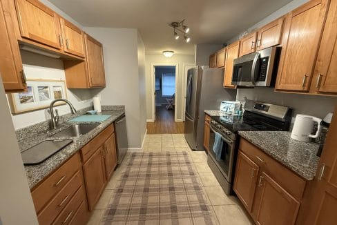 Galley kitchen with wooden cabinets, granite countertops, stainless steel appliances, and a plaid rug on a tiled floor, leading to a dining area with wood flooring.