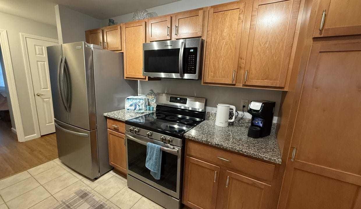 A modern kitchen with stainless steel appliances, wooden cabinets, a gas stove, microwave, refrigerator, and countertop with a coffee maker, water bottles, and paper towels.