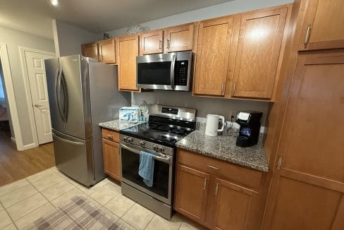 A modern kitchen with stainless steel appliances, wooden cabinets, a gas stove, microwave, refrigerator, and countertop with a coffee maker, water bottles, and paper towels.