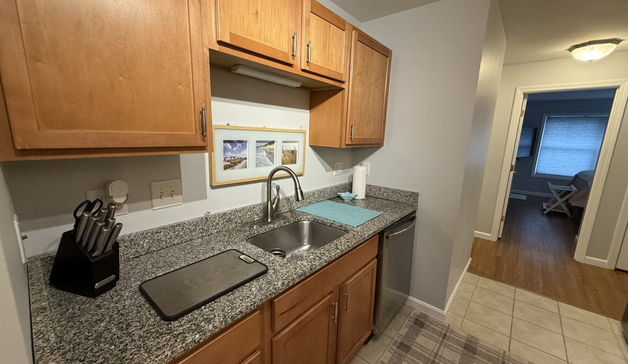 Small kitchen with wood cabinets, granite countertop, sink, knife block, cutting board, and a blue drying mat; hallway leads to a room with a window and wooden floor.