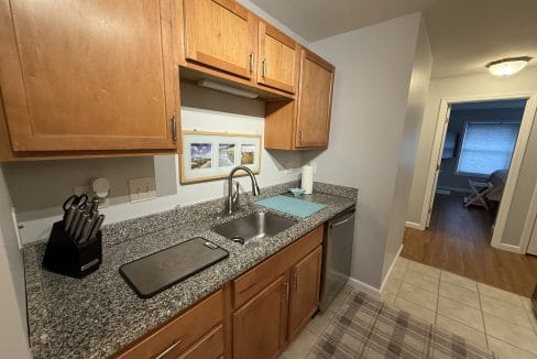 Small kitchen with wood cabinets, granite countertop, sink, knife block, cutting board, and a blue drying mat; hallway leads to a room with a window and wooden floor.