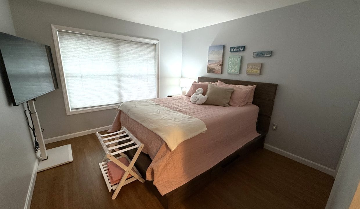 A bedroom with a large bed, pink bedding, wall art, a TV mounted on the wall, wooden floors, and a window with blinds letting in natural light.