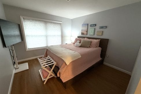 A bedroom with a large bed, pink bedding, wall art, a TV mounted on the wall, wooden floors, and a window with blinds letting in natural light.