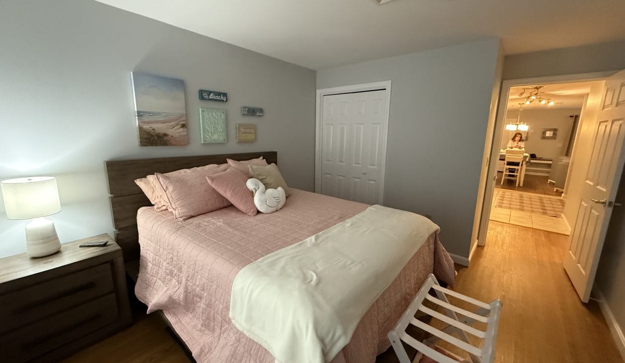 Bedroom with a queen bed, pink bedding, two bedside tables, a lamp, wall art above the headboard, a folded towel on the bed, and a view into a hallway with dining area.